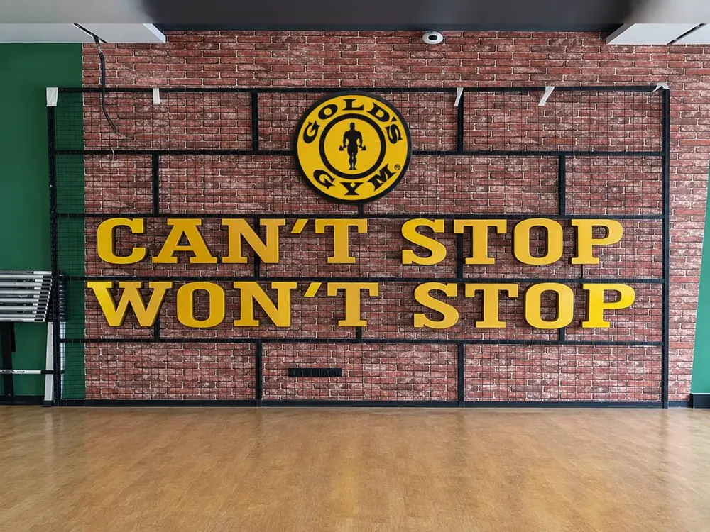 Gold's Gym interior brick wall with black metal grid, circular Gold's Gym logo and large yellow 3D letters reading Can't Stop Won't Stop above a wooden floor